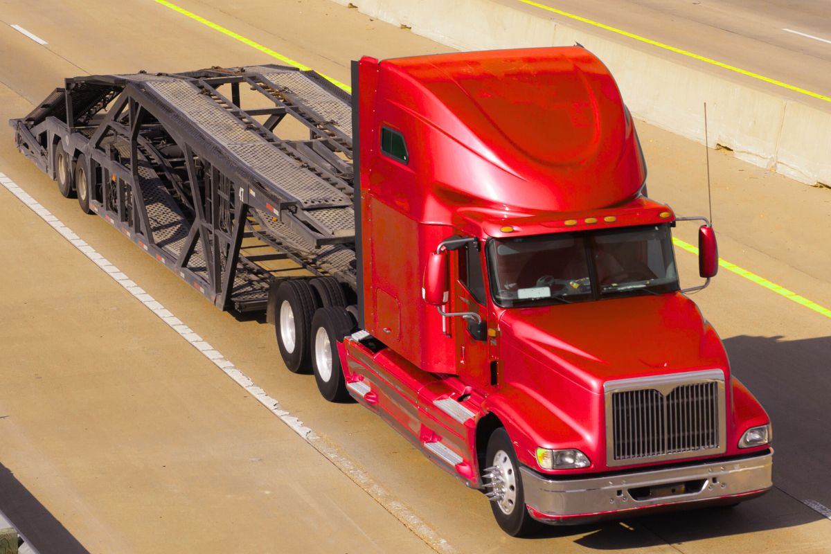 Red open auto transport truck driving on highway, used for vehicle shipping across long distances.